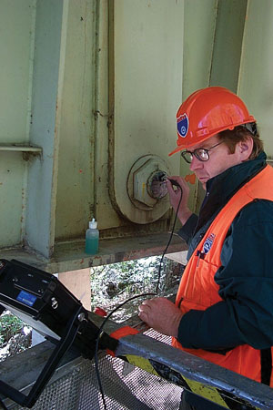 A worker in safety glasses, helmet, and vest is using the Ultrasonic bridge evaluation system on a metal structure.