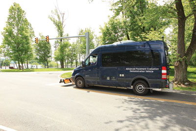 Partial side view of a van at a demonstration intersection with equipment mounted on the front bumper, rear wheel, and top. The van is labeled "Advanced Equipment Evaluation Demonstration Vehicle."