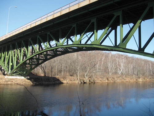A side view of a single span of a steel-truss deck bridge is shown from ground level, crossing a river. Bare trees are shown on the opposite shore.