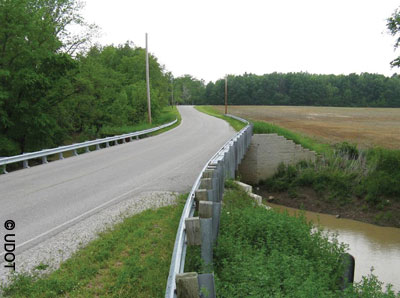 View of a two-lane bridge with guardrail over a narrow creek in a rural area. Farmland is on one side and a wooded area on the other.