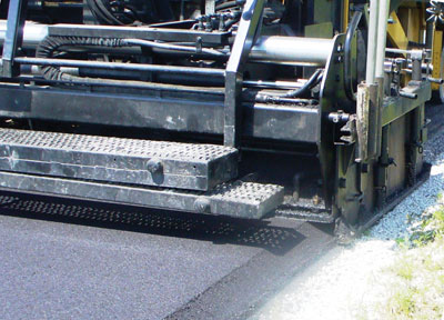 Photo. The rear view of an asphalt paving machine as it consolidates the edge of a pavement to 30 degrees to create the "Safety Edge."