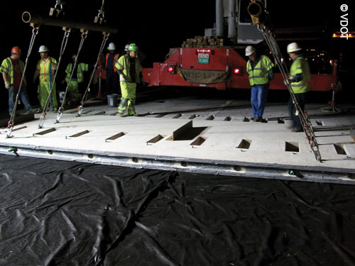 Night view of placement of precast, prestressed concrete panels in a roadway. In the foreground is sheeting covering the base. A long panel is being supported using heavy equipment  visible in the background. Workers in safety gear are looking on.
