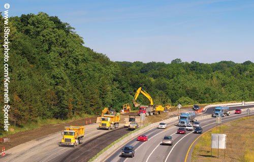 Overhead view of a highway construction scene, with four lanes of traffic approaching on the right and construction taking place left of the median. Three dump trucks are visible, including one being loaded by a shovel, a bulldozer, and other construction equipment.