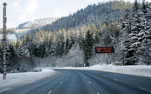 A winter scene showing four clear lanes of curved highway in one direction with snowy forested hills ahead. Plowed snow is a few feet high on either side of the roadway. A variable message sign indicates "Speed Limit 55," "Snow slush and ice in places," and a temperature of 25 degrees.