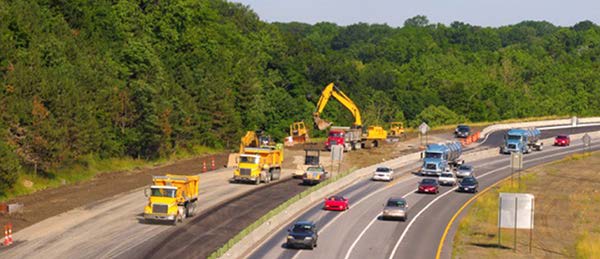 Stock photo shows a highway from a distance. Several lanes of the highway are blocked off, with construction vehicles on them. Three lanes on the right side of the photo have normal highway traffic.