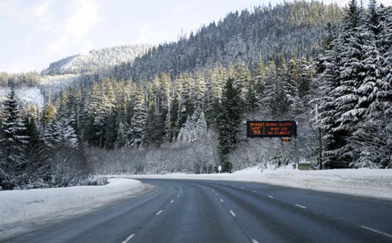A stock photo shows an empty highway in a hilly, wooded area. An electronic sign alongside the road includes information about highway conditions and speed limits.