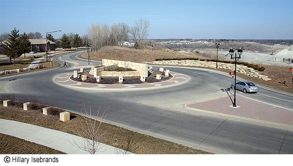 Coralville, IA Roundabout Isebrands FHWA.jpg. Figure 1. Photo. A roundabout in a rural area with roadways entering from the top, bottom, and left sides of the photo. A raised brickwork median separates traffic lanes where the roadways enter the circle. On the left side of the roadway is a sidewalk, and on the right side there is a low stone wall with shrubbery behind it. In the center of the circle is a landscaped area containing a low stone wall in the shape of a circle dissected by a taller stone wall in the direction of the roadway that enters and leaves the circle; and at right angles, a lower wall pointing to the roadway that completes the junction. Just outside this structure are white cube-shaped markers with black arrows pointing clockwise (the direction of traffic flow). Outside this central area is a wide patterned brickwork circle incorporating white arrow heads that also point counterclockwise. A car is entering the circle in the foreground. On the other side of the circle a pedestrian is crossing the road. Four sets of streetlights are visible. Houses and trees appear in the distance.
