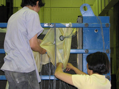This photo shows two workers carefully pulling the strain gauge cables through the membrane sheet and around parts of the test bin.