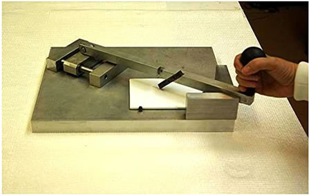 Figure 3. Photo. Example of a scribing tool. This photo shows a scribing tool that was designed and built at the Coatings and Corrosion Laboratory. The tool is made of stainless steel consisting of a 14.5-by-11-by-1-inch (368.3-by-279.4-by-25.4-mm) support base to rest the test panel while mechanically scribing it. The test panel is held in place by raised hinges. The surface of the base consists of two fixtures: a guide rod fixture and a v-notch. A handle consisting of a scribing tip fixed at an angle slides back and forth on support guide rods to scribe the test panel. When the handle is not in motion, it rests in the v-notch. 
