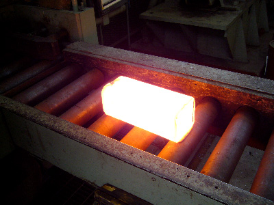 This photo shows a bright, nearly white, rectangular block of steel on a roller table on the entry side of a reversing rolling mill. The block is 5 inches (127 mm) thick and 11 inches (279 mm) long.
