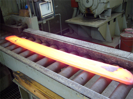 This photo was taken on the entry side of a reversing plate mill and shows a plate approximately 5 by 1 by 50 inches (127 by 25.4 by 1,270 mm). The color of the steel is yellow, indicating a temperature close to 1,800 °F (982 °C), except for a region on the right end of the top surface of the plate that is gray, indicating a temperature below 1,200 °F (649 °C). This is a surface defect that developed during the hot rolling operation.