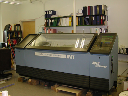 The photo shows a cyclic corrosion chamber from the front. It is approximately 10 by 4 by 5 ft (3 by 1.2 by 1.5 m) and has three major sectional elements. To the left is a reservoir for solutions that are periodically sprayed on the samples located beneath the transparent cover in the center section. On the right side of the chamber is the control section.