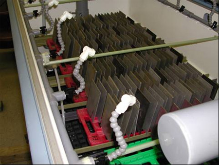 This photo shows the interior of a cyclic corrosion chamber. It is taken from above the open cyclic corrosion chamber and shows many 4- by 6-inch (100 - by 150-mm) corrosion test panels arranged in a near-vertical orientation, each separated from its neighbor by about 0.5 inches (12.7 mm). Also shown are spray bar headers along the front and rear edges of the inside of the test chamber from which adjustable piping lead to white spray heads directed downward onto the test panels.