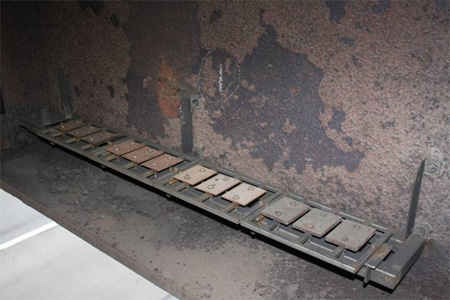 This photo shows corrosion coupons on Moore Drive Bridge on rack 1 just prior to removal after 1 year of exposure. The photo was taken from above, and the I-390 southbound lane is visible in the lower left corner. The upper half of the photo shows the web of a bridge girder that is heavily corroded. Bolted to the web is a rack oriented horizontally about 2 inches (50.8 mm) above the bottom flange of the girder, also shown to be heavily corroded. The specimen rack contains four sets of three coupons, each held in place with two bolts.