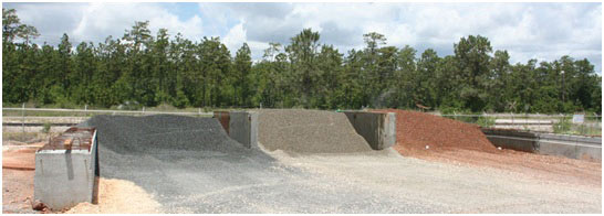 Figure 1. Photo. Lightweight aggregate stockpiles with continuous sprinklers. This photo shows three separate piles of lightweight aggregate. A small garden sprinkler on top of each aggregate pile is spraying water into the air above each pile.