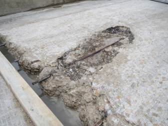 Figure 8. Photo. Distressed region on the westbound lane near the western abutment: test region B1.(2) This figure shows a photo of the distressed region located on the westbound lane near the west abutment. A large, oval-shaped region of distressed concrete can be observed. Within the oval-shaped region, a single corroded steel reinforcing bar is present. The photo also shows that there is some deterioration along the west abutment line of the bridge structure.