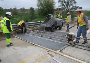 Figure 10. Photo. UHPC overlay installation on the westbound lane.(2) This figure shows a photo of the UHPC overlay material being placed on the westbound lane of the bridge. This was the first of the two installation phases. UHPC is being placed using a motorized buggy. Workers are assisting with placement using hand tools. The overlay is being finished using a large vibrating screed that is manually moved by workers.