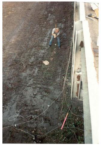 SASW tests at bent 12, structure 4, Trinity River Relief Bridge. This image shows tests conducted at bent 12 of relief structure 4 of the Trinity River Relief Bridge using the spectral analysis of surface wave method. It shows a worker digging beneath the bridge, with wires hanging from the bridge.