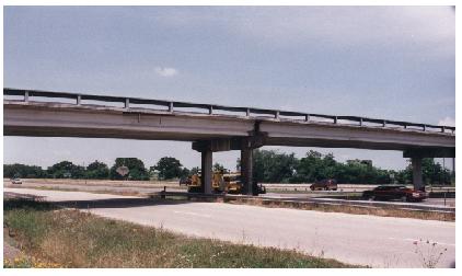 Western pier of the southwestern span of the Woodville Road Bridge. This image shows the western pier of the southwestern span of the Woodville Road Bridge in Bryan, Texas. The photo was taken from the grassy shoulder of Texas State Highway 6 looking forward and slightly up at the bridge. The photo shows three of the four prestressed concrete I girders resting directly on the beam caps through polystyrene bearing pads. A truck used for dynamic testing is parked under the bridge.