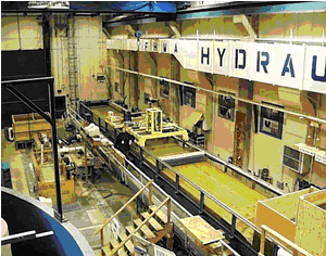 Figure 1. View of the flume in the Hydraulics Laboratory. Photo. This picture shows an overhead view of the Hydraulics Lab at the Turner-Fairbank Highway Research Center in McLean, Virginia. The flume is on the right side of the picture. It is elevated above the lab floor, and there is a walkway next to the flume that is accessible via a set of stairs in the foreground.