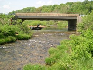 Figure 14. Meander migration affecting right abutment, Hammond Branch, Maryland. Photo. This figure shows a river flowing toward a bridge. A meander bend upstream of the bridge turned within about 30.5 meters of the bridge.