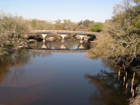 Figure 129. Peace River, Atlantic Coastal Plain-upstream from bridge at old pedestrian bridge. Photo. This is the Peace River in the Atlantic Coastal Plain region looking upstream from the bridge toward a pedestrian bridge upstream. The left bank is heavily vegetated with trees, and the right bank is sparsely vegetated. 