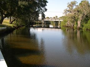 Figure 134. Alligator Creek, Atlantic Coastal Plain-downstream from bridge. Photo. This is the Alligator Creek in the Atlantic Coastal Plain region looking downstream from the bridge. The left bank has only grass and is rigidly stabilized with a stone wall. The right bank is very heavily vegetated with trees. 