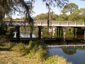 Figure 135. Alligator Creek, Atlantic Coastal Plain-looking downstream at bridge. Photo. This is looking downstream toward the bridge. The channel is well-aligned with the bridge, and the flow is very calm. The left bank is grassed and stabilized with a stone wall. The right bank is lined with trees.
