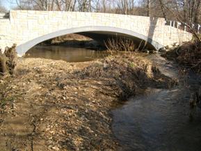 Figure 212. Indian Run, Piedmont-looking upstream at bridge. Photo. This is looking upstream at the bridge. The large central bar causes an obstruction to flow, forcing the flow into the opposite bank.