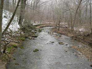 Figure 221. Little Elk Creek, Piedmont-upstream from bridge. Photo. This is the Little Elk Creek in the Piedmont region looking upstream from the bridge toward a meander bend. Bank vegetation consists of moderately dense trees. There is a narrow gravel bar along the left bank. 