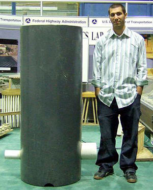 Figure 4. Photo. Access hole prototype in the 1986-1992 study with the lab technician to show the relative size. This picture shows a man approximately 8.3 meters (6 feet) tall standing next to a PVC model of an access hole, which is about as tall as the top of his shoulders and is a little wider than his body. Near the bottom of the access hole model is a small PVC inlet pipe on the left and a larger PVC outlet pipe on the right.