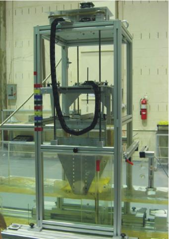 Figure 13. Photo. Deck force analyzer system at the TFHRC hydraulics lab. This vertical format photograph shows the deck force analyzer as mounted in a flume filled halfway with water. The flume crosses the bottom of the picture horizontally. A tall frame, roughly square in plan view, straddles the flume. The test section is connected to the top of the frame by long, straight pendulums. The test section has two identical sections of the type shown in figure 12 sitting along the side walls of the flume. The flume is roughly half filled with water, and the test section holds the streamlined bridge deck just above the water level.