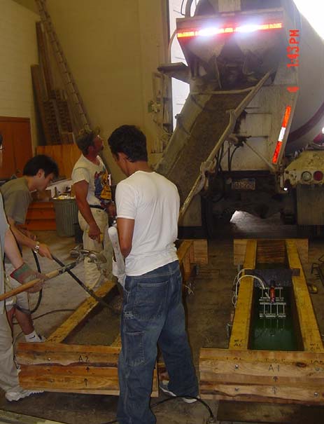 This photo shows ready-mixed concrete being unloaded from the mixing truck through chutes and poured into the wooden formwork. Three people are spreading and vibrating the concrete mix in the formwork.