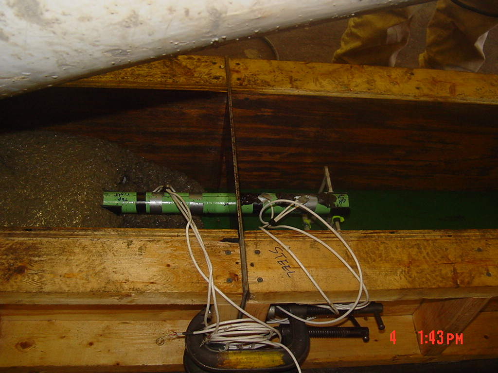 This photo shows a dowel bar with strain gauge and wire attachment placed in the wooden formwork as it is being covered with the concrete pouring down from the chute of a concrete mixer. 