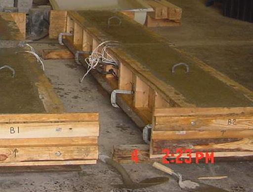 This photo shows surface-finished jointed concrete pavement slabs just after they are cast in wood formwork. Hooks are provided on both sides across the joints, and strain gauge wires coming out of the slab from the dowel bars are visible.