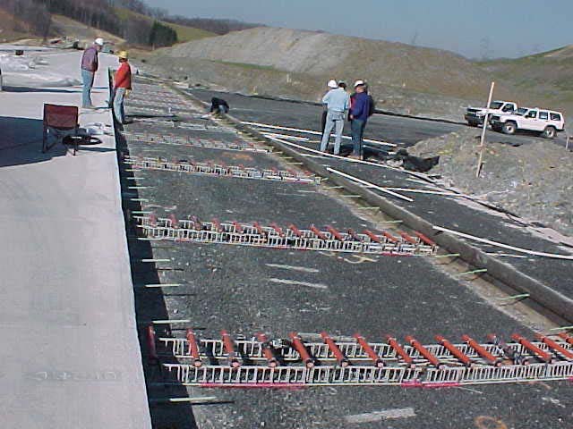 This photo shows field installation of several rows of fiber reinforced polymer (FRP) dowel bars on baskets at location 2 of corridor H project on Route 219, Elkins, WV. Plastic pipes are shown on the shoulder side of the pavement which carries strain gauge wires from the instrumented dowels to outside of the shoulder region. Personnel involved in the project are also seen.