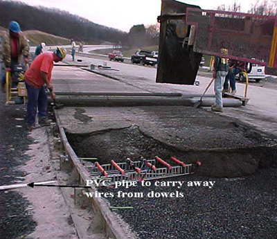 This photo shows part of the construction truck and construction crew participating in the construction of jointed plain concrete pavement. Fiber reinforced polymer (FRP) dowel bars are being covered by the concrete and a vibrator is being used for concrete compaction. Polyvinyl chloride pipes carrying the wires from the instrumented dowel bars are also seen.
