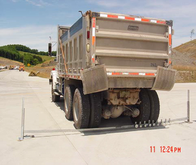 This photo shows a rear view of a West Virginia Department of Transportation truck with calibrated loads. The truck is positioned at a concrete pavement joint with instrumented fiber reinforced polymer dowels. Slab deflections caused by the truck wheel load are being measured by a series of dial gauges suspended from a long metal frame resting on adjacent lanes.