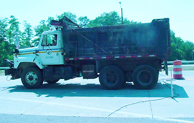 This photo shows a side view of the West Virginia Department of Transportation (WVDOT) truck used for field test loading on the concrete pavement joint with fiber reinforced polymer dowels. A metal stand assembly supporting dial gauges and the linear variable differential transformer including the wire running from the linear variable differential transformer are also seen.
