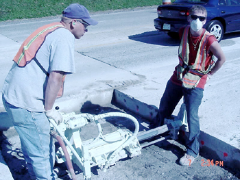 This photo shows holes being drilled by two persons on the side of the concrete pavement slab cut to insert fiber reinforced polymer dowel bars during the rehabilitation process. 