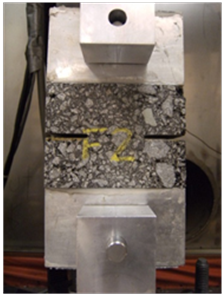 This photo shows a double-edged notched tension (DENT) specimen for essential work of fracture (EWF) and critical tip opening displacement (CTOD) characterization. A rectangular block of asphalt mix with two notches cut on each side in the middle is glued between two aluminum end caps that have been mounted in two clevises and pins at each end.