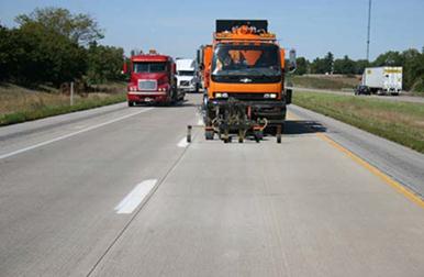 This photo shows a truck applying pavement marking material in the left lane of a roadway while traffic passes it in the right lane.