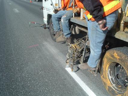 This photo shows a truck sprayer with two workers monitoring the equipment as paint is applied on a roadway.
