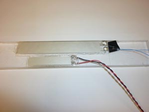 This photo shows a polyvinylidene fluoride (PVDF) piezo film bounded to a Plexiglas® beam. There are two rectangular metallic objects (the PVDF piezo film), one of which is smaller and is attached to a clear beam of Plexiglas®. There are wires extending from each object from the right side.
