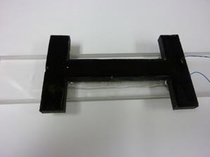 This photo shows the polyvinylidene fluoride (PVDF) embedded in epoxy and bounded to the Plexiglas® beam. There is a black object in the shape of a horizontally stretched  H  attached to a clear beam. Extending from the right of the H-shaped object (i.e., the PVDF embedded in epoxy)are two wires.