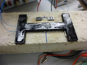 This photo shows a polyvinylidene fluoride (PVDF) piezo film embedded in epoxy and then bound to a concrete slab. A black H-shaped PVDF piezo film is attached to the concrete slab. There are two blue wires extending from the PVDF.