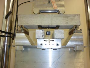 This photo shows the bending test setup to check the activation strain loss. The photo shows the concrete slab with the polyvinylidene fluoride piezo film embedded in epoxy attached to it. The concrete slab is in the testing apparatus face down, and each end of the concrete is supported by a roller. The part of the apparatus that applies the load is above the concrete.