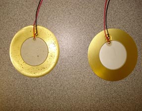This photo shows a piezoelectric transducer covered with a layer of rubber. Two circular disks are shown; the inside is white, and a gold colored ring surrounds it. There are two wires extending from each disk.