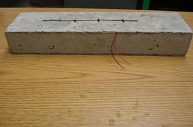 This photo shows a concrete specimen with an embedded piezoelectric generator. The concrete block is shown with a red and black wire extending from the top corner.