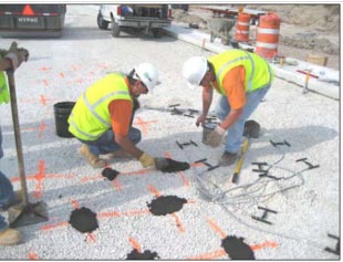 This photo shows two construction workers placing sand/binder pads and fitting gauges on the orange markings on the ground.